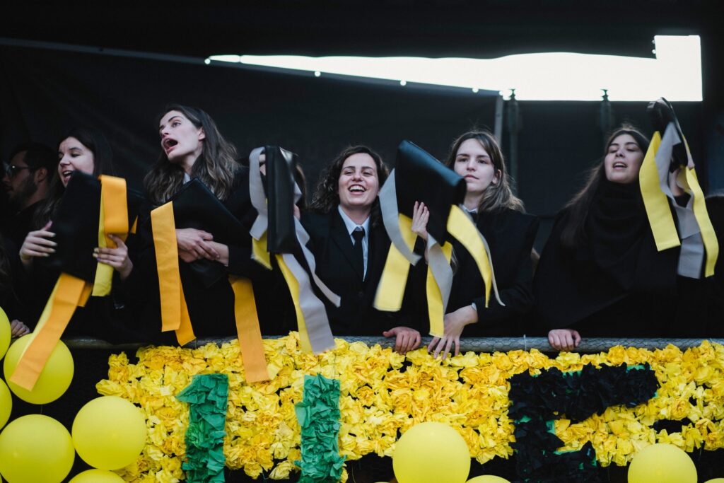 Fotografija prikazuje študente, ki sodelujejo v edinstveni univerzitetni tradiciji, z nasmejanimi obrazi in simboli kulturne dediščine posamezne univerze.